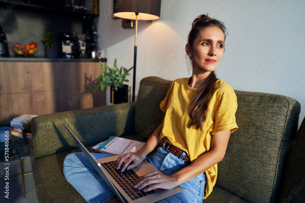 Fototapeta premium Young caucasian woman sitting on sofa and working on laptop while looking away
