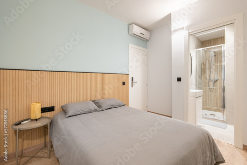 Bedroom with spacious bed against wall demarcated with wood paneling and blue paint and bedside table. Bedding is in beautiful gray color. From room there is doorway to bathroom with glass shower. © Pavel
