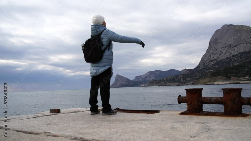 A woman tourist raises her hands up, standing on a pier above the sea against the background of the sky and mountains, dressed in a blue jacket and black trousers with a black backpack. Observation of A woman tourist raises her hands up, standing on a pier above the sea against the background of the sky and mountains, dressed in a blue jacket and black trousers with a black backpack. Observation of
