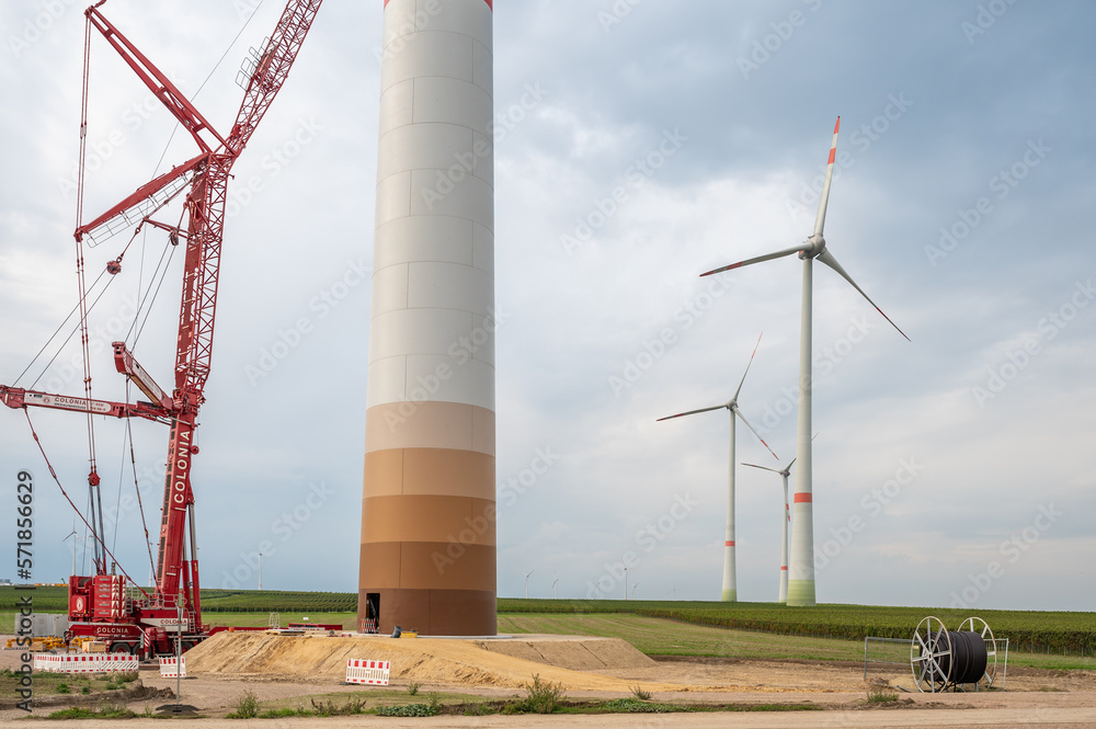 Construction site of a wind turbine, crane next to the tower of the wind turbine, completed wind turbines in background, cable reel, low angle view, cloudy day Construction site of a wind turbine, crane next to the tower of the wind turbine, completed wind turbines in background, cable reel, low angle view, cloudy day
