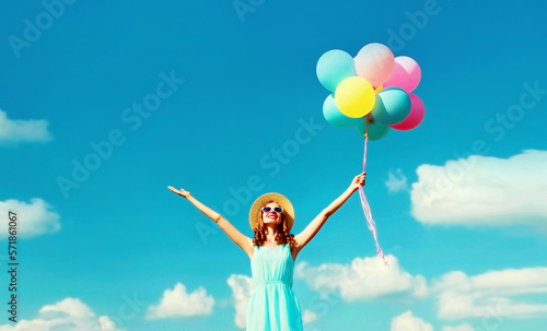 Happy smiling young woman with bunch of colorful balloons wearing summer straw hat on the field on blue sky clouds background © rohappy