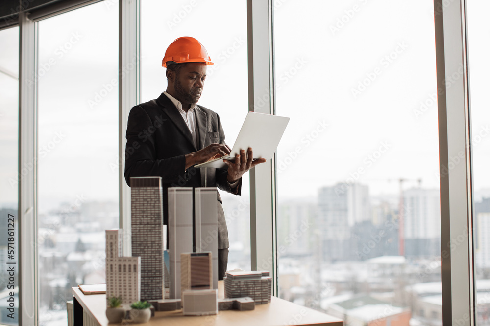 Handsome chief architect african man in business suit and protective helmet with laptop pc working with city buildings model as urban planner at office interior with panoramic city view. Handsome chief architect african man in business suit and protective helmet with laptop pc working with city buildings model as urban planner at office interior with panoramic city view.