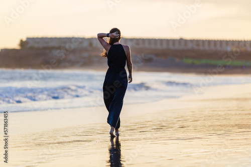 Young beautiful woman in long blue dress walking at la tejita beach at sunset. Young lady in long dress enjoying the evening at the beach photo