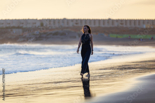 Young beautiful woman in long blue dress walking at la tejita beach at sunset photo