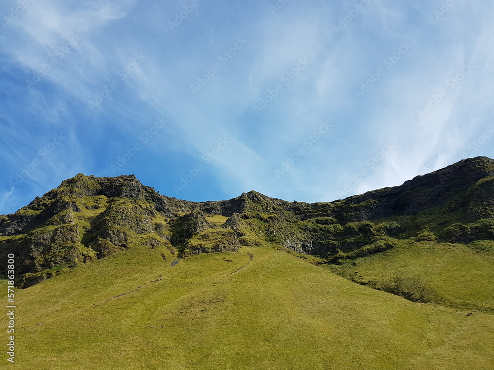 Grass-covered hill with blue sky and thin white clouds in Iceland Grass-covered hill with blue sky and thin white clouds in Iceland