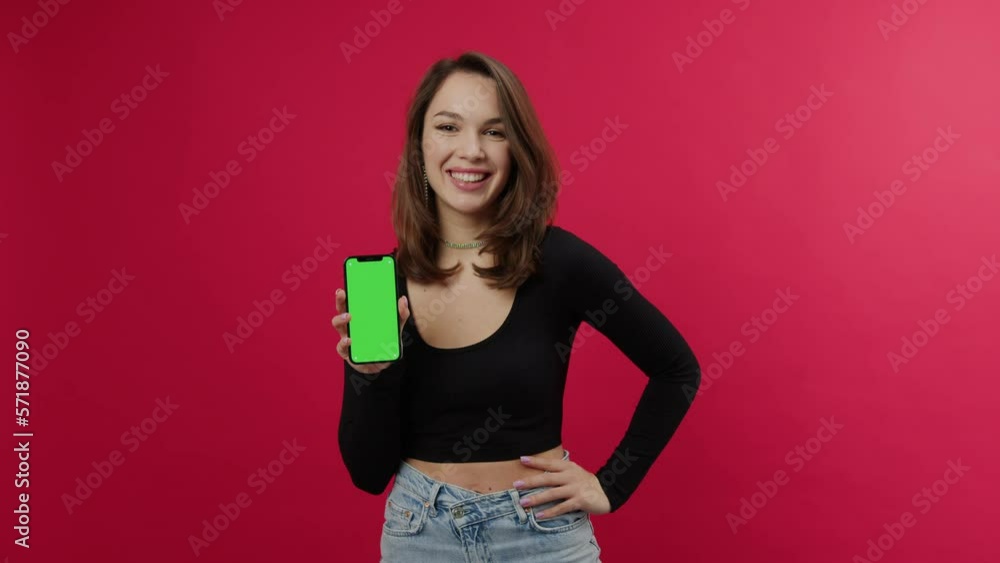 A young woman with a smile picks up a mobile phone with a green screen. The model looks at the camera on a red background. Placing ads on the gadget A young woman with a smile picks up a mobile phone with a green screen. The model looks at the camera on a red background. Placing ads on the gadget