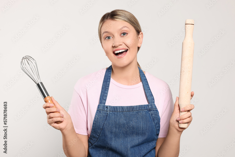 Beautiful young woman in clean apron with kitchen tools on light grey background Beautiful young woman in clean apron with kitchen tools on light grey background