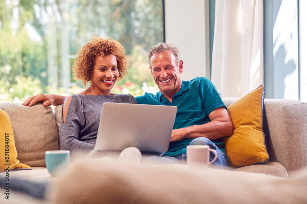 Senior Retired Couple Sitting On Sofa At Home Shopping Or Booking Holiday On Laptop Senior Retired Couple Sitting On Sofa At Home Shopping Or Booking Holiday On Laptop