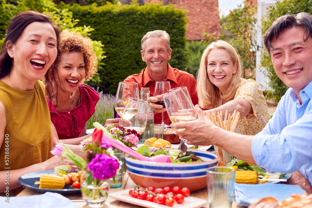 Portrait Of Mature Friends Around Outdoor Table Making A Toast With Wine At Summer Party In Garden 