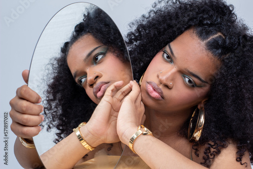 Mujer joven latina con el pelo rizado y un vestido de tirantes de varios colores asomándose por detrás de un espejo redondo con su reflejo en él. photo