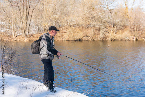 A fisherman with a fishing rod and a backpack catches fish on the bank of a snow-covered river