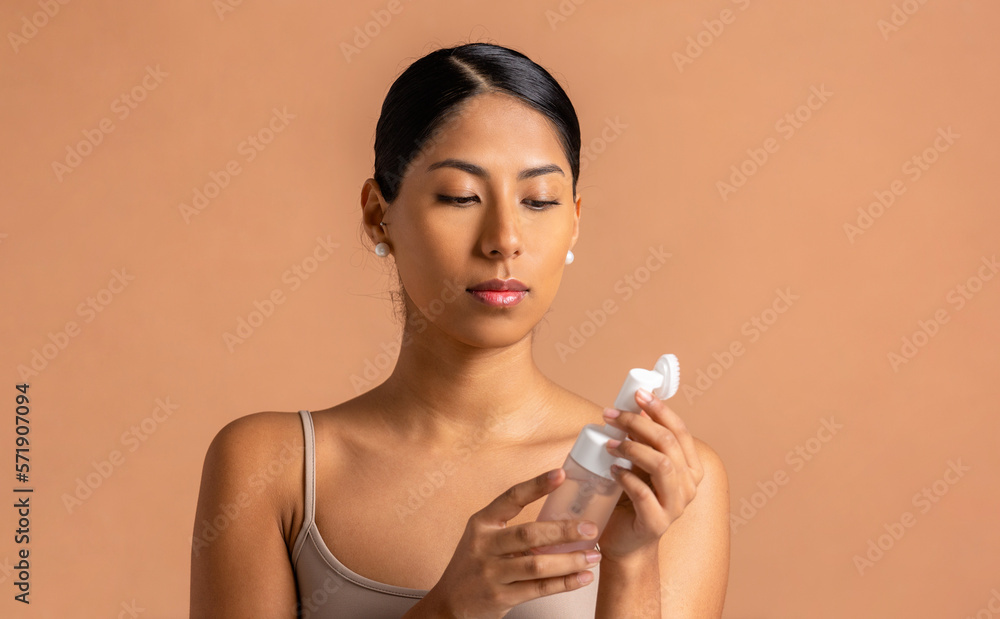 Portrait of young ethnic woman with glass bottle of aromatic essential oil for spa session and facial massager against beige background Portrait of young ethnic woman with glass bottle of aromatic essential oil for spa session and facial massager against beige background