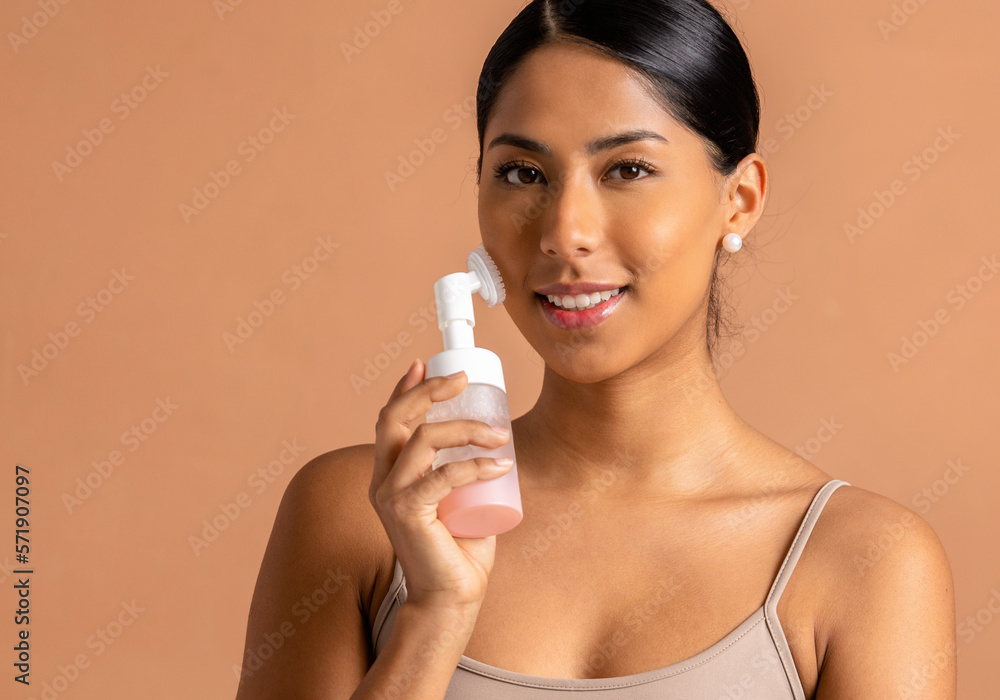 Portrait of young ethnic woman with glass bottle of aromatic essential oil for spa session and facial massager against beige background Portrait of young ethnic woman with glass bottle of aromatic essential oil for spa session and facial massager against beige background