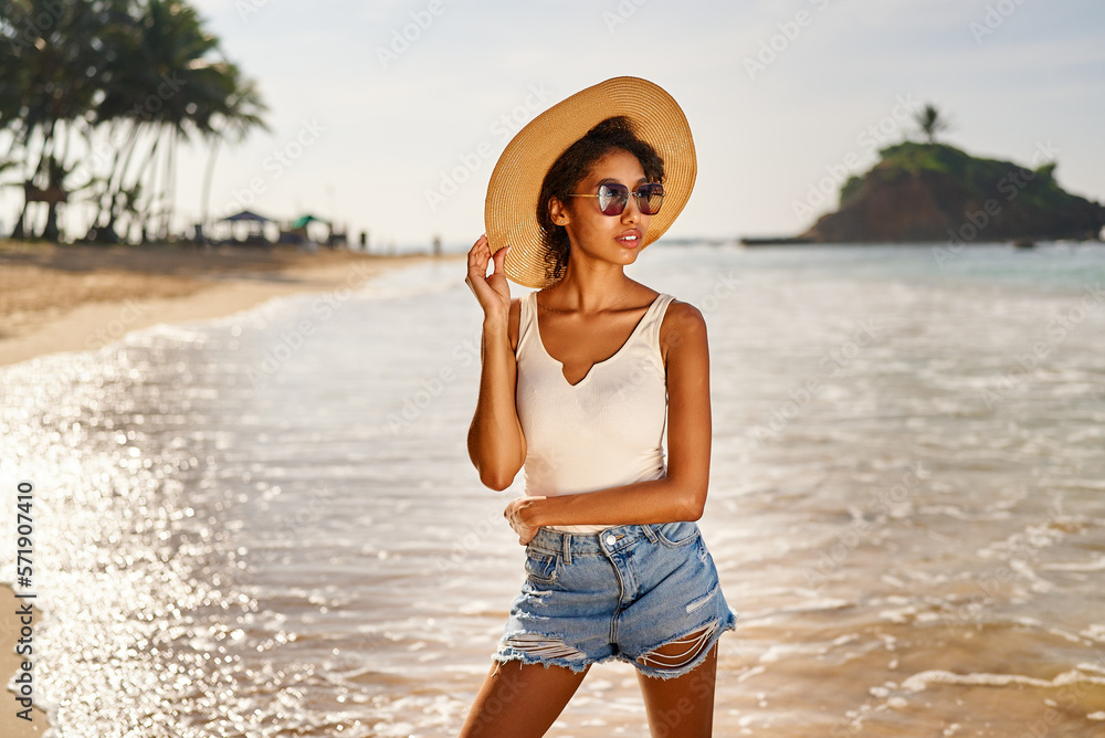 Young african female model in straw hat and sunglasses posing at resort by sea at sunrise. Black woman against scenic rocky green island and ocean tide at dawn. Multiracial model poses on vacation