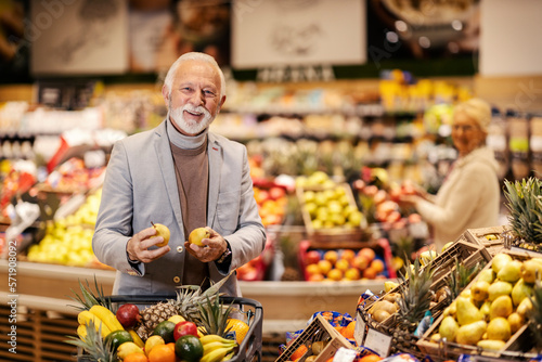 A happy old man is filling his shopping cart with fresh fruits at the supermarket while smiling at the camera.