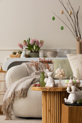 Warm and cozy composition of easter living room interior with coffee table, white sofa, colorful easter eggs, easter bunny, hyacinth, vase with branch and personal accessories. Home decor. Template.
