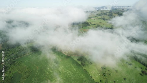 Aerial above the clouds over beautiful green fields, pasture and a river seen through holes in the clouds. Drone. Low clouds. 4K. 