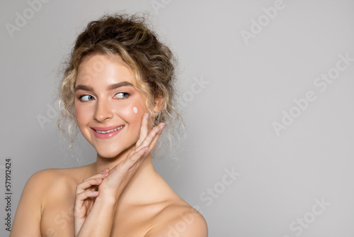 Face skin care. Attractive lady applying cosmetic cream on clean hydrated skin, looking aside at free space