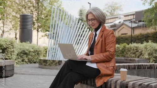 Senior attractive Woman using laptop in front of Business building. Pretty grey hair elderly female sitting on the bench in a city park with coffee and work online