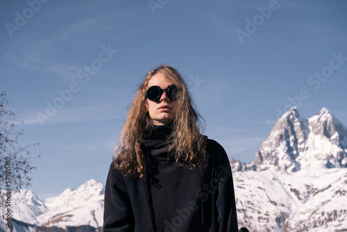 Portrait of stylish man wearing round sunglasses in front of snowy mountain peaks on vacation