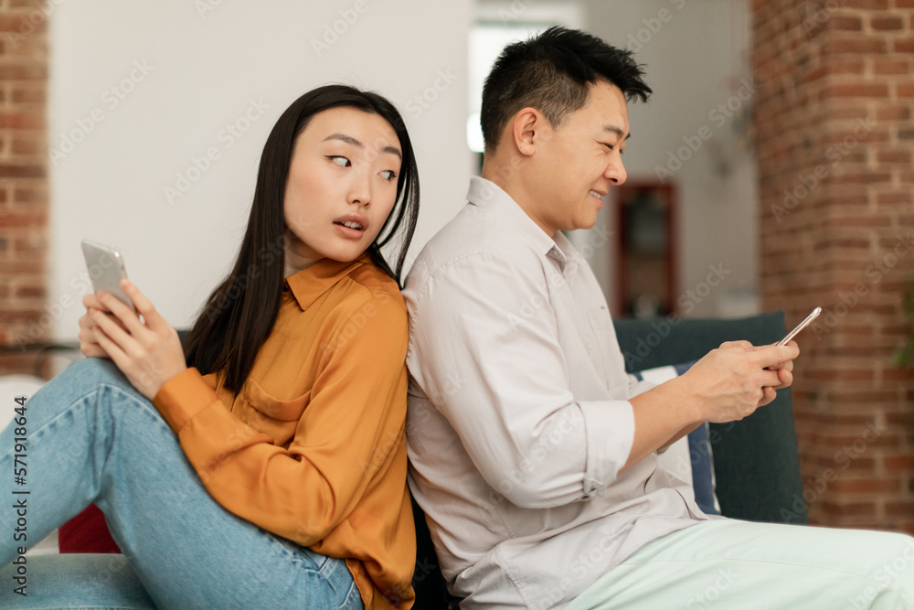 Jealousy concept. Jealous asian womam reading husband's messages on cellphone while man texting and smiling Jealousy concept. Jealous asian womam reading husband's messages on cellphone while man texting and smiling