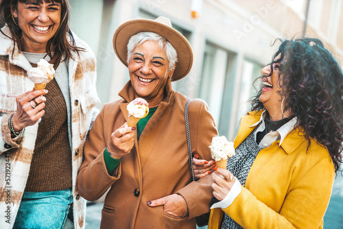 Three mature women eating ice cream cone outside - Older female friends having fun walking on city street - Joyful elderly lifestyle concept photo