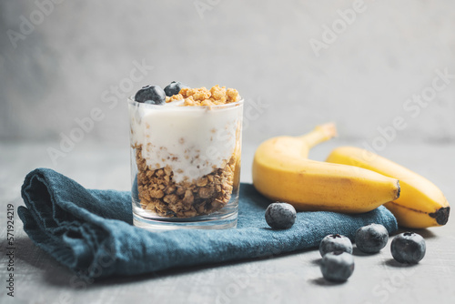 Granola cereal oatmeal with white yogurt, blueberries and banana fruits in a glass on a blue napkin, grey background