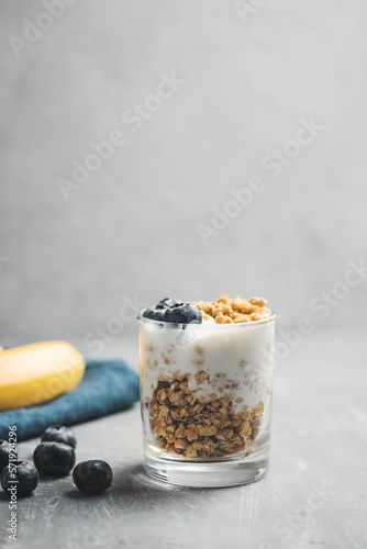Granola cereal oatmeal with white yogurt, blueberries and banana fruits in a glass on a grey background