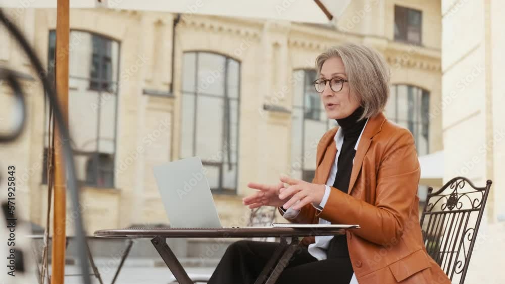 mature gray-haired business woman sitting outdoors in a cafe holds an online conference with a laptop mature gray-haired business woman sitting outdoors in a cafe holds an online conference with a laptop