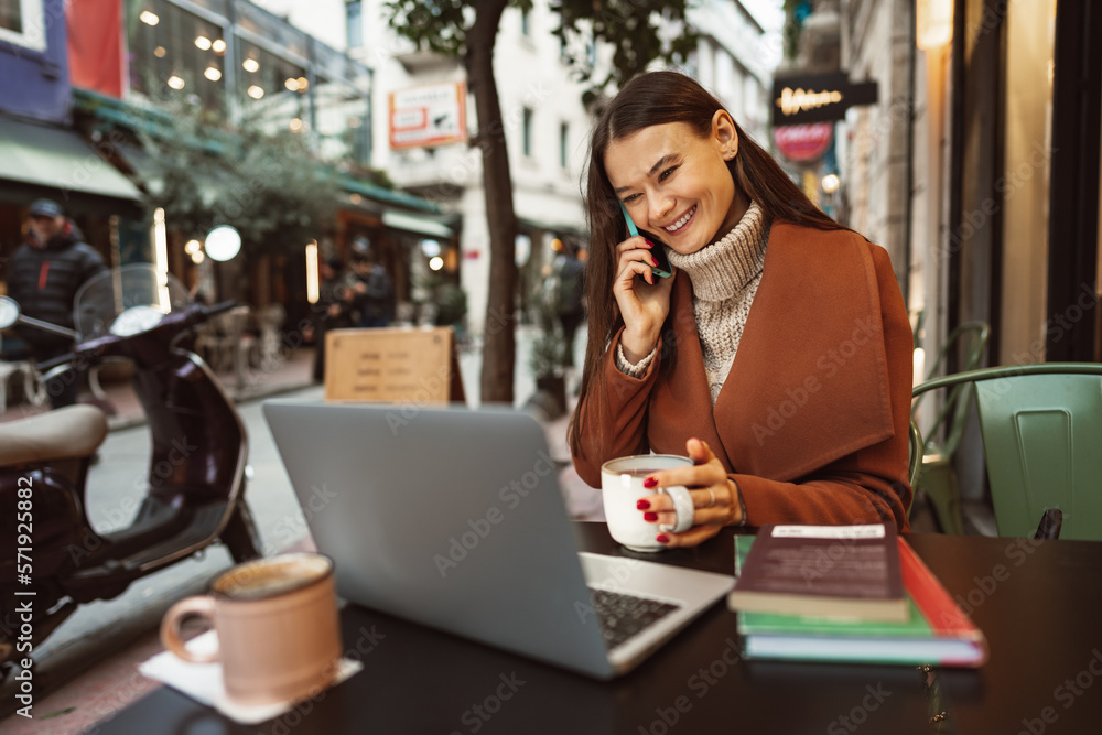 Smiling young woman using laptop and and talking on the phone while sitting in cafe