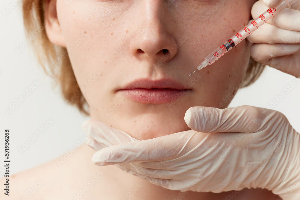 Young redhead man having cosmetological injections. Male model posing against grey background. Lip augmentation. Concept of men's health, cosmetology, plastic surgery, skin care, hygiene, medicine