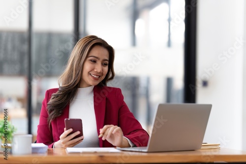 Smiling beautiful Asian businesswoman analyzing chart and graph showing changes on the market and holding smartphone at office