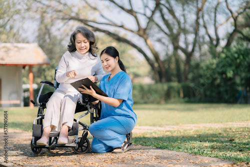 young woman in blue workwear nurse helping senior woman in wheelchair with questionnaire, asian pensioneer filling papers at nursing home, having assistance.