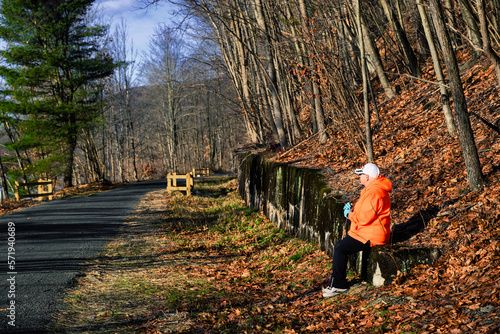 My wife takes a rest while we are walking along the D&H Rail Trail in Lanesboro, PA this Winter. A woman in a bright orange coat and white hat sits along the trail enjoying a beautiful winter's day. photo