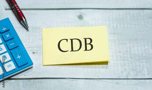 A sigla CDB de Certificado de Depósito Bancário escrita em um pedaço de papel que está sobre uma mesa de madeira. Uma calculadora e uma caneta na composição. Economia brasileira e investimentos. photo