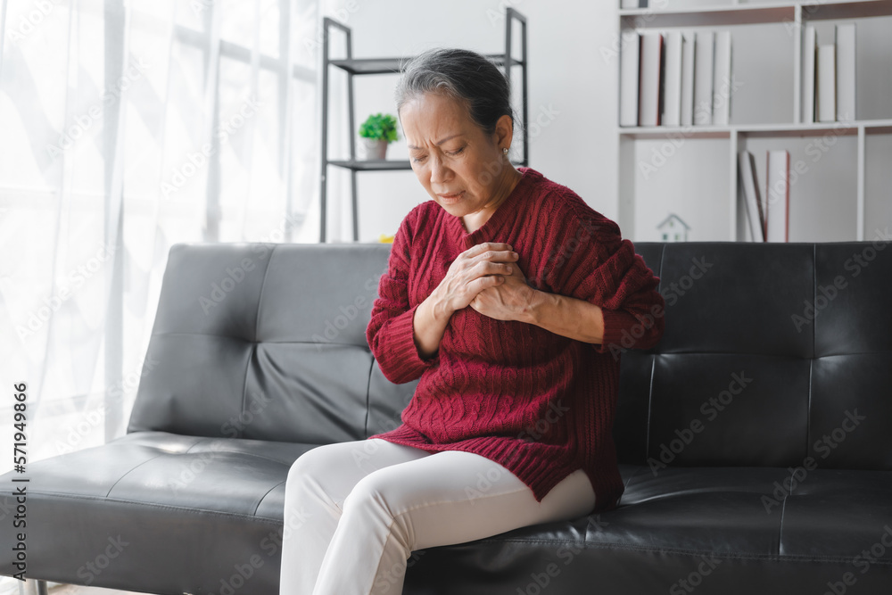 Elderly asian housewife woman sitting on sofa. Heart disease, chest pain, irregular heartbeat Elderly asian housewife woman sitting on sofa. Heart disease, chest pain, irregular heartbeat