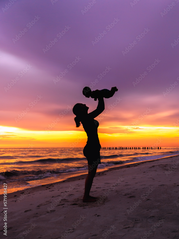 Young adult woman mother holding baby in air on beach with sunset background silhouette, vertical empty space