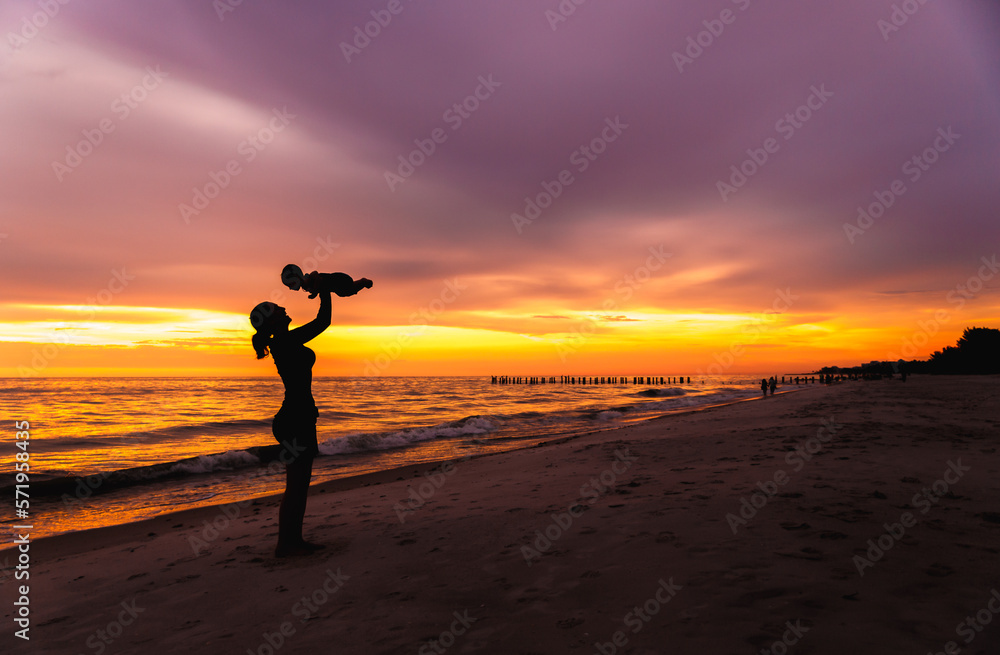 Young adult woman mother holding baby in air on beach with sunset background silhouette, horizontal empty space