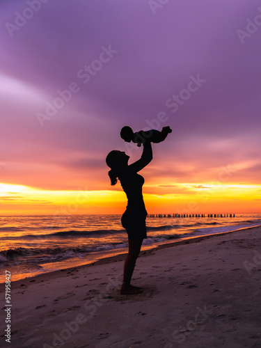 Young adult woman mother holding baby in air on beach with sunset background silhouette  vertical empty space