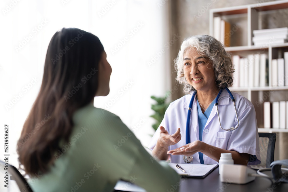 Woman senior doctor is Reading Medical History of Female Patient and Speaking with Her During Consultation in a Health Clinic. Physician in Lab Coat Sitting Behind a Laptop in Hospital Office