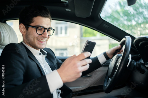 A successful businessman is a driver in a car, a man writing a message on the phone, using an application for motorists, a person in a positive mood. © muse studio