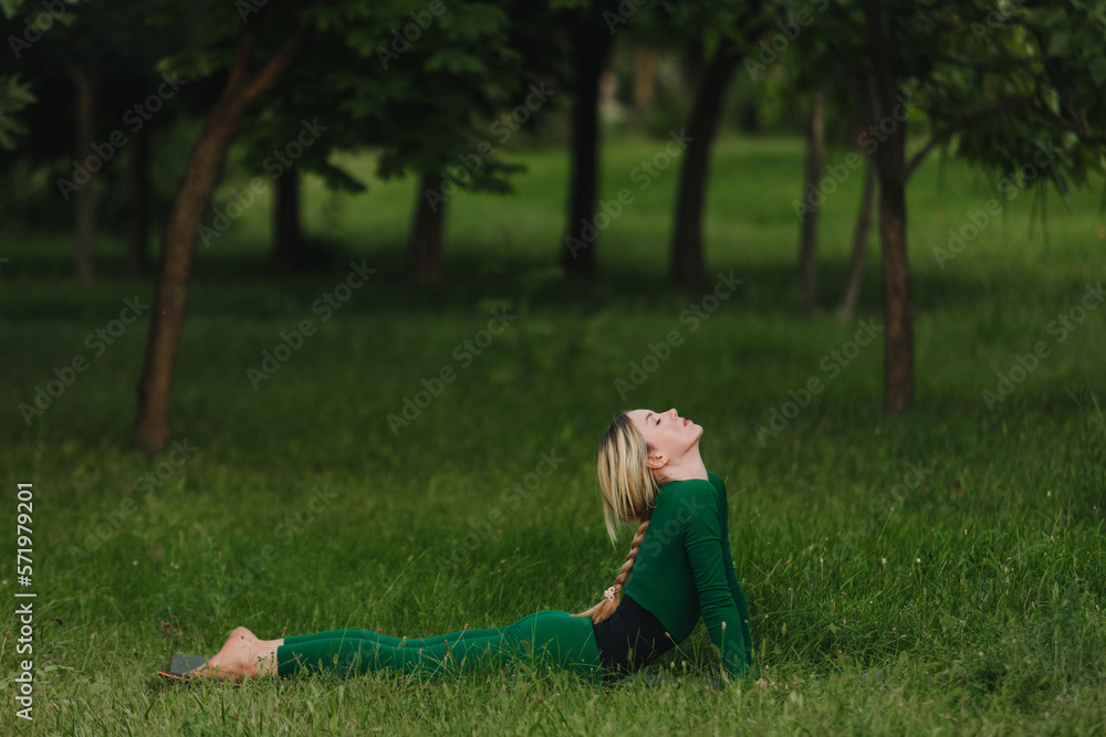 Fototapeta premium Young woman doing cobra pose on yoga mat in the park on a body summer evening.