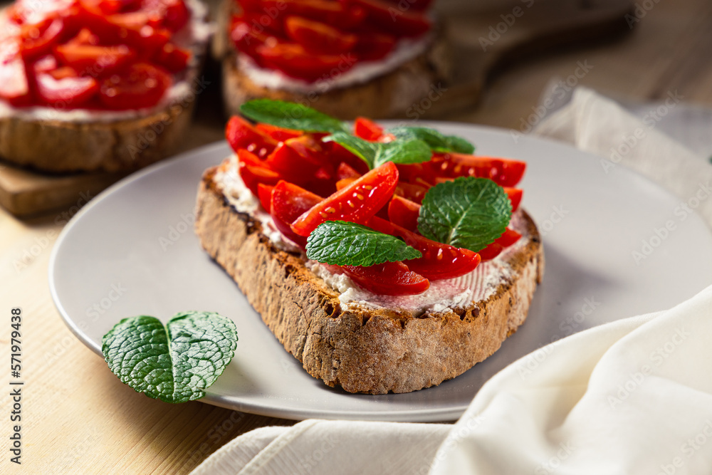 Front view of three pieces of bruschetta on a plate with cherry tomatoes, cream cheese and mint leaves on a wooden table
