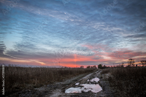 The field road between the villages is covered with puddles that reflect the colors of the dawn sun.