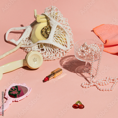 Rotary dial phone in a mesh bag, lipstick, retro style hand mirror, pearls, coupe champagne glass, creative aesthetic layout, pastel pink background. Nostalgia trend. 