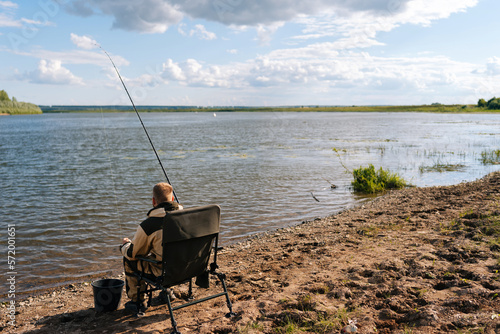 Rear view of unrecognizable fisherman wearing raincoat sitting on river bank on travel chair with fishing rod catches fish on summer sunny day. Concept of lifestyle, leisure activity on nature