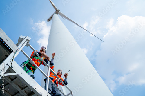 Lower view of two engineers or technician workers stand on base of big windmill or wind turbine and point or look to left side with blue sky.