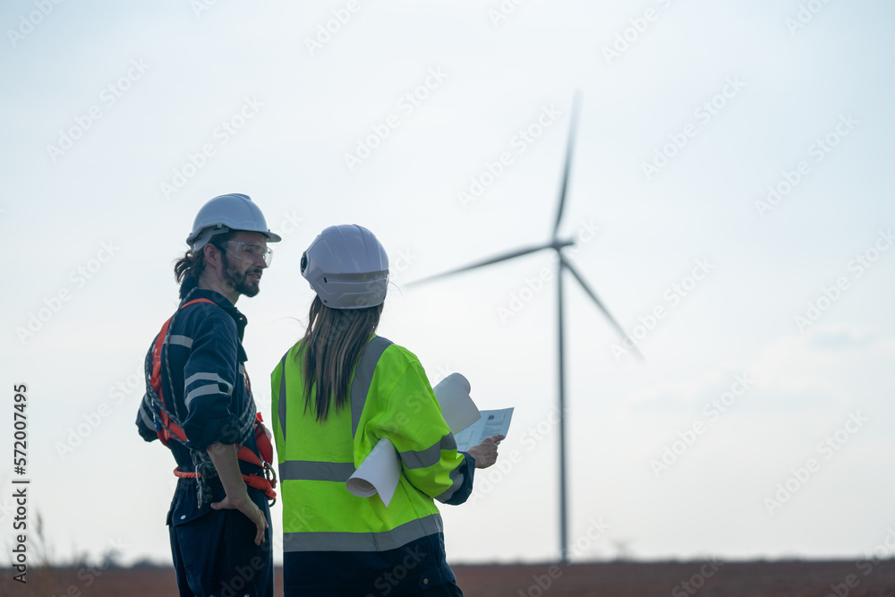 Back of Caucasian engineer man and woman discuss together with woman hold drawing paper and stay in front of row of windmill or wind turbine with blue sky. Back of Caucasian engineer man and woman discuss together with woman hold drawing paper and stay in front of row of windmill or wind turbine with blue sky.