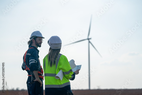 Back of Caucasian engineer man and woman discuss together with woman hold drawing paper and stay in front of row of windmill or wind turbine with blue sky.