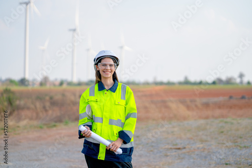 Pretty engineer or technician woman worker hold drawing paper and look at camera with smiling also stand in front of windmill cluster in workplace.